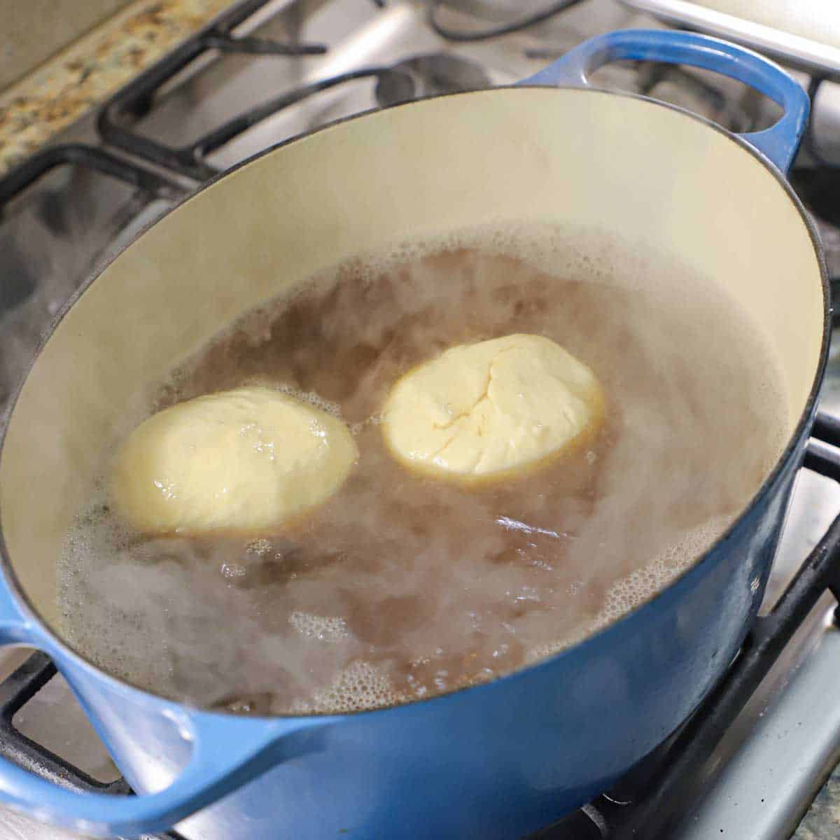 Two balls of pretzel bun dough in boiling water and molasses in a large oval Dutch oven on a gas stove.