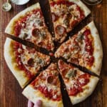 A person pulling a slice of homemade New York-style pizza from the sliced pie that is sitting uneaten on a wooden cutting board.