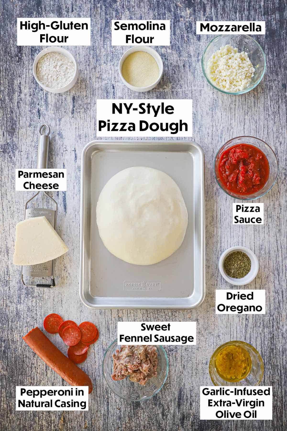 An arrangement of ingredients for homemade New York-style pizza on a grey wooden background including flour, cheese, dough, pizza sauce, pepperoni, sausage, and seasonings.