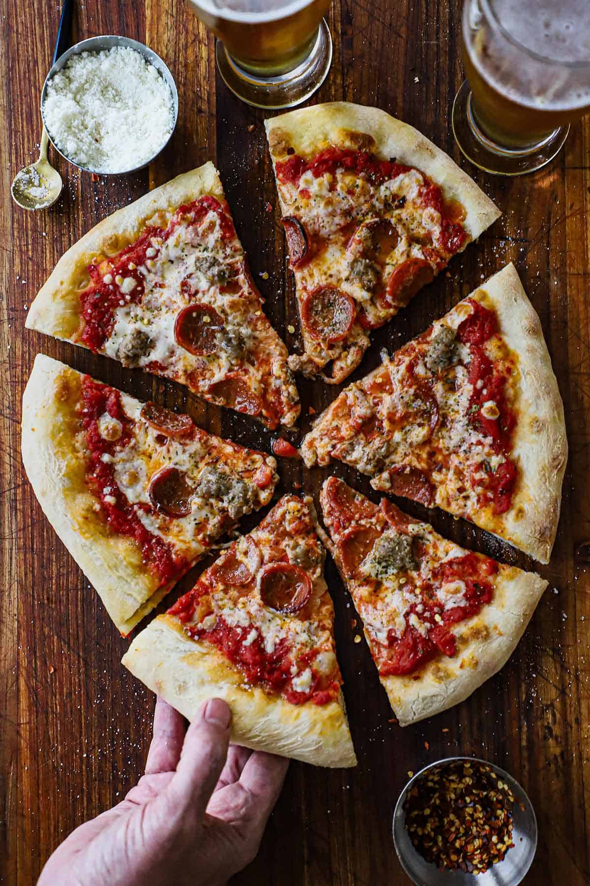 A person pulling a slice of homemade New York-style pizza from the entire pie that has been cut into six slices with a small bowl of grated cheese and two glasses of beer nearby.