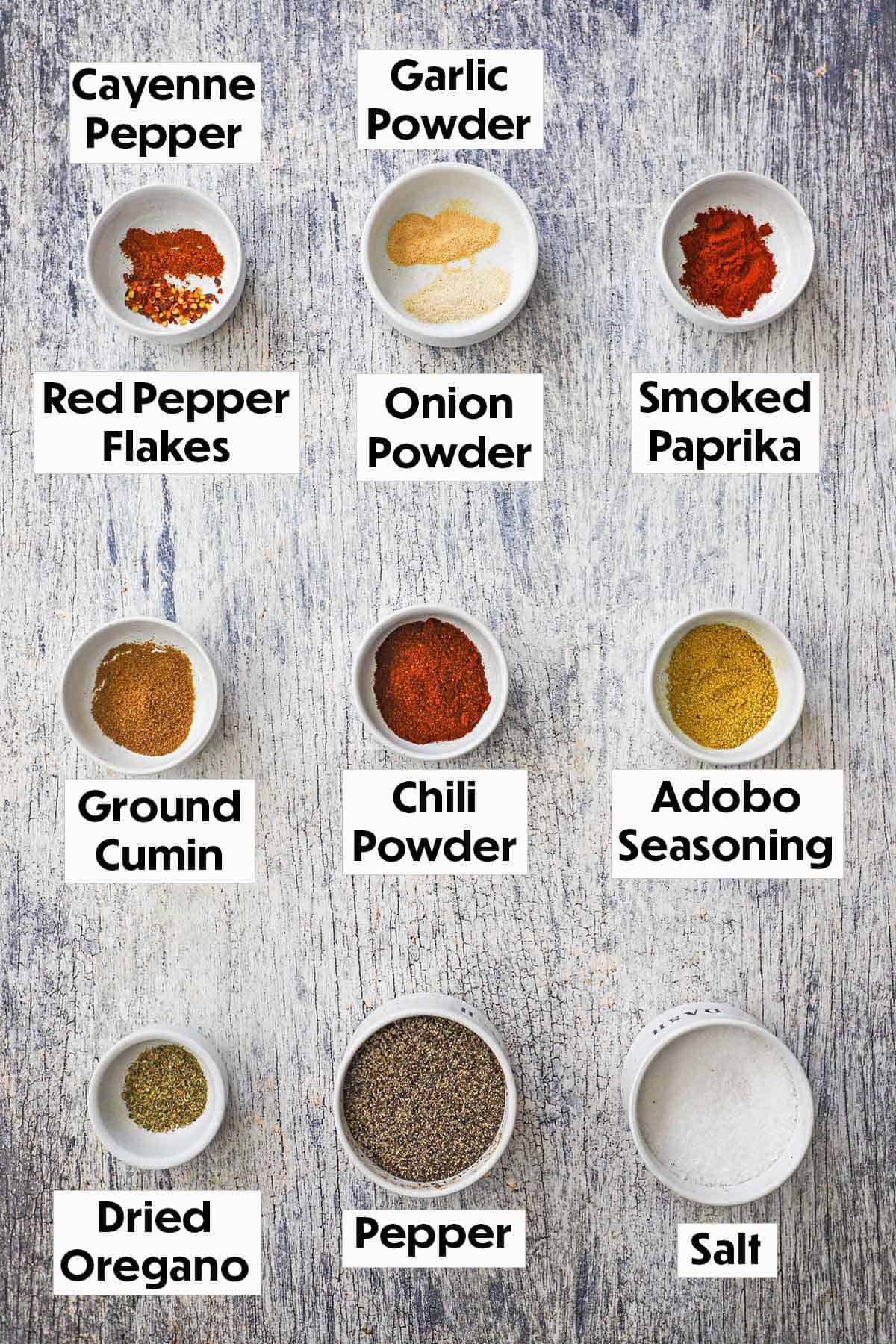A arrangement of small white bowls filled with seasonings for homemade taco seasoning on a grey wooden background including chili powder, paprika, cumin, adobo seasoning, garlic powder, oregano, salt, and pepper.