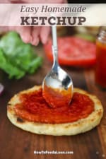 A person using a small spoon to smear easy homemade ketchup all over the top of a toasted hamburger bun on a wooden cutting board.
