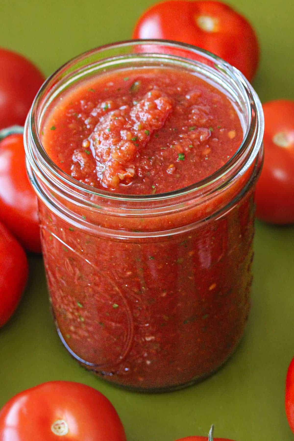 A clear jar that is filled with restaurant-style salsa on a green background and is surrounded by fresh whole tomatoes.