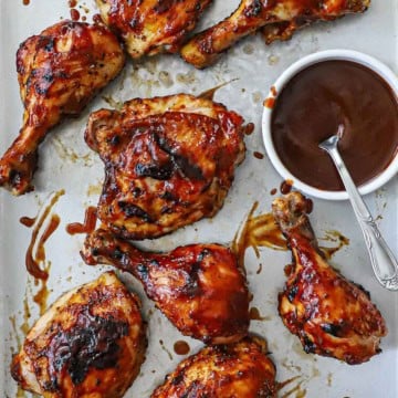 Grilled BBQ Chicken with visible grill marks resting on a metal sheet pan with a small white bowl of homemade BBQ sauce with a spoon in it nearby.