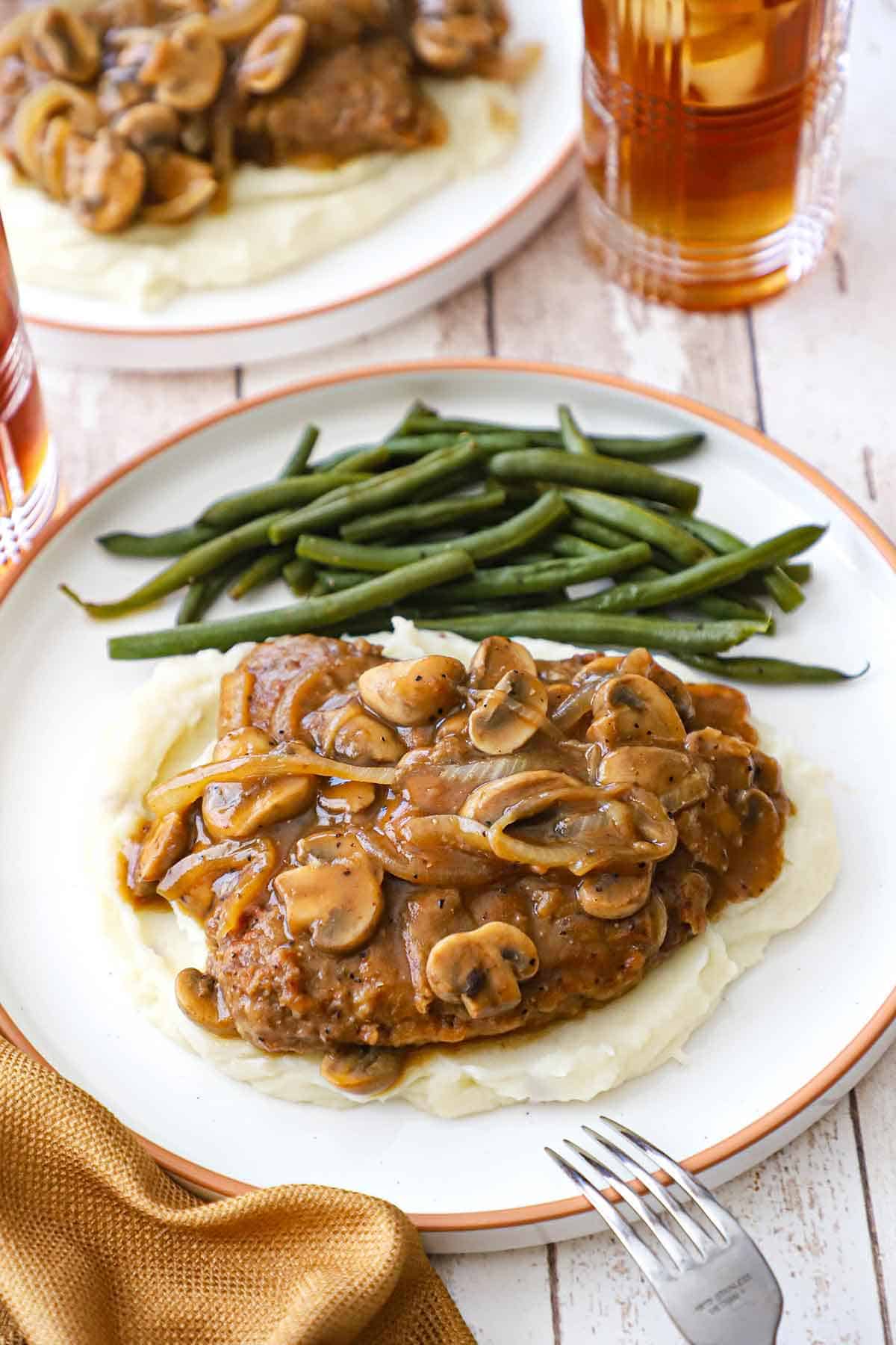 A white dinner plate filled with a serving of cube steak with mushroom gravy resting on a bed of mashed potatoes with braised green beans off to the side.