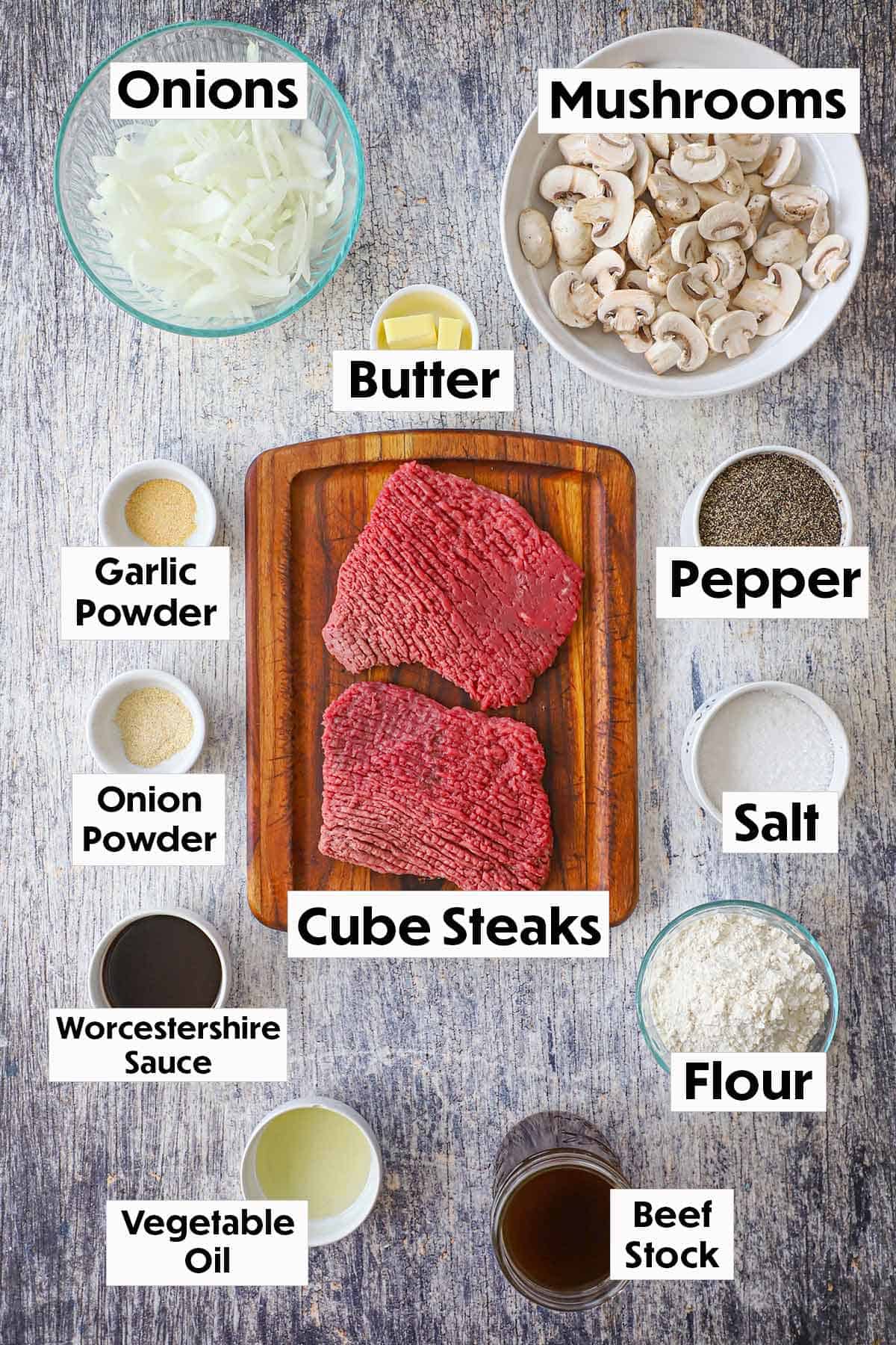 An arrangement of ingredients for cube steak with mushroom gravy on a grey wooden background including cube steak, onions, mushrooms, Worcestershire sauce, flour, and seasonings. 