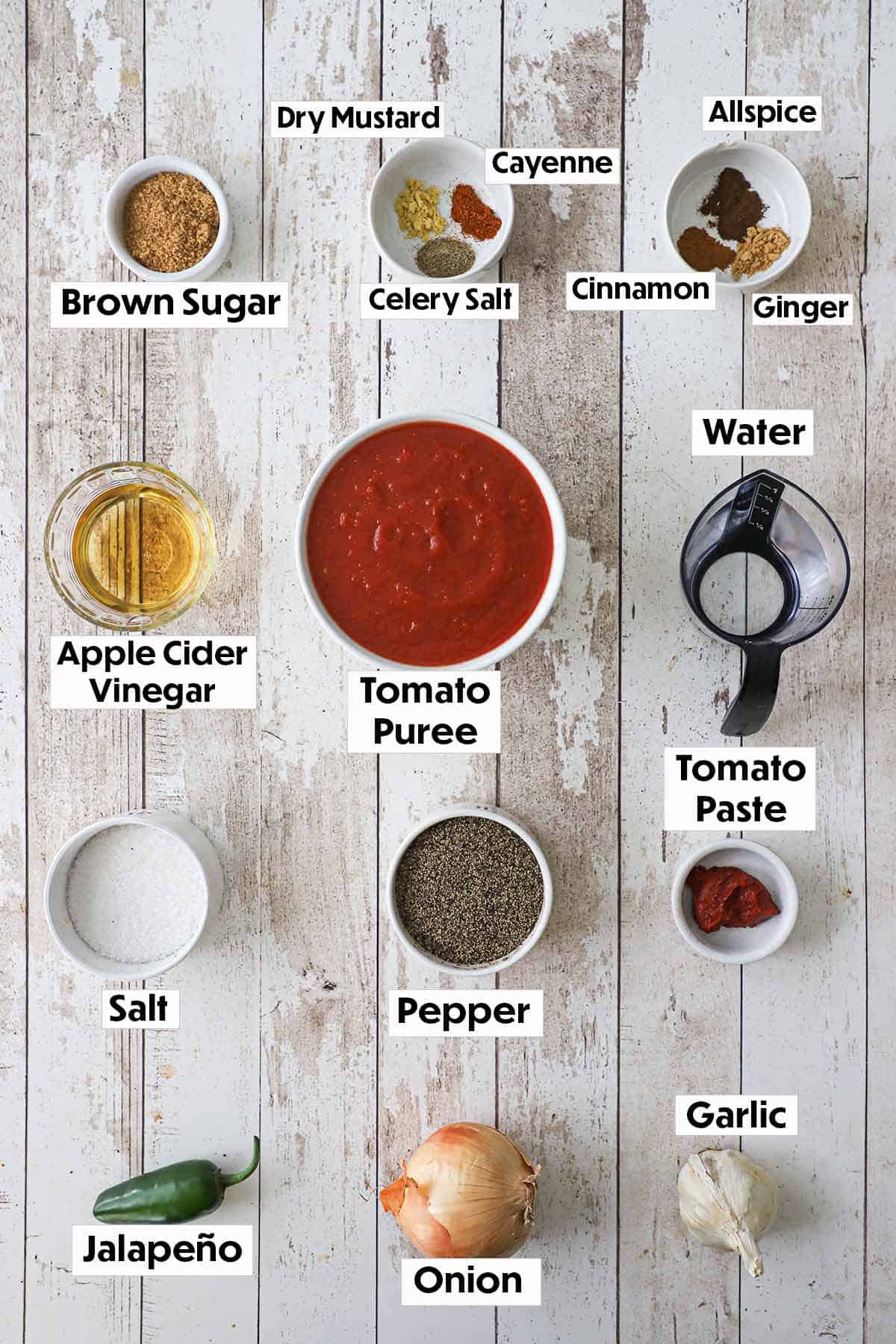 An arrangement of ingredients for easy homemade ketchup on a grey wooden background including bowls of tomato purée, brown sugar, seasonings, vinegar, water, tomato paste, jalapeño, onion, and garlic.