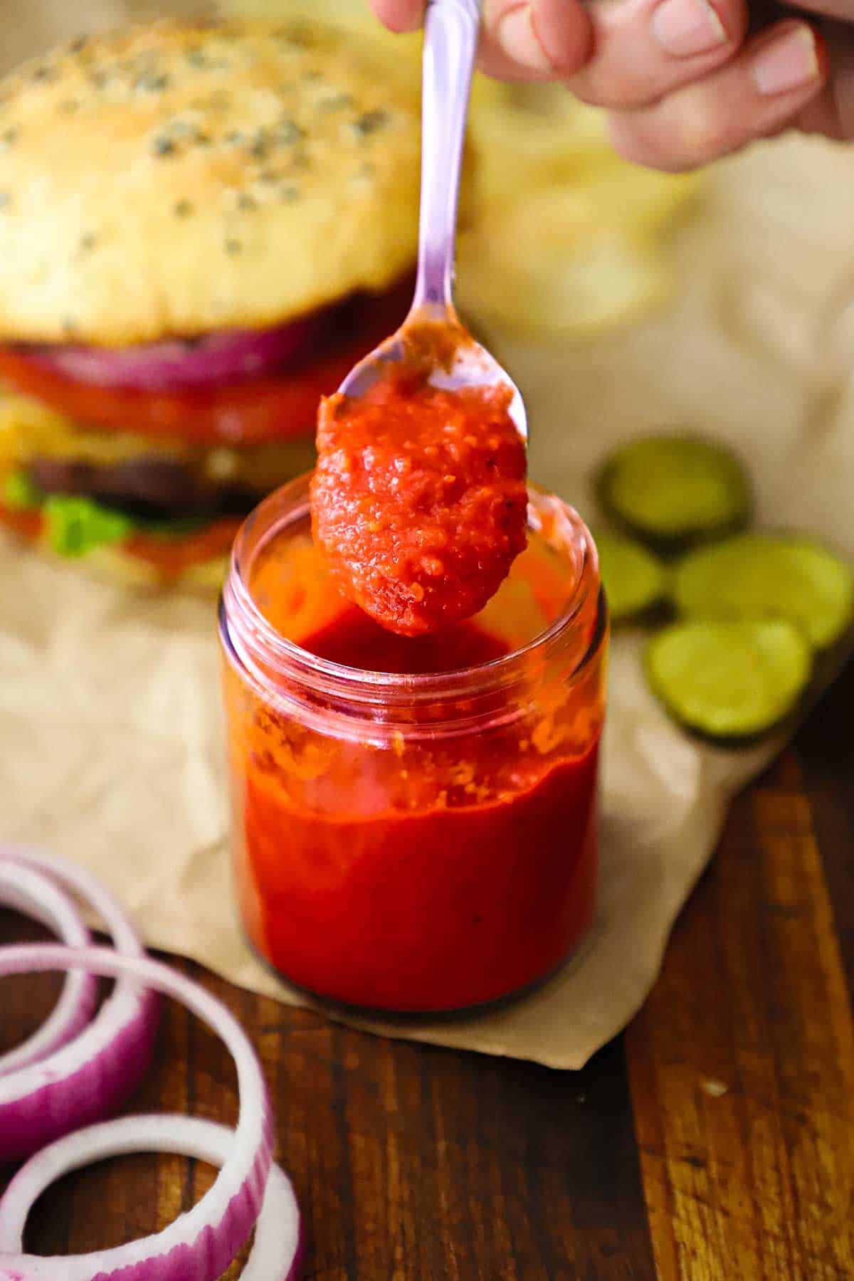 A person holding up a spoonful of easy homemade ketchup over a small glass jar of the same next to a cheeseburger resting on a crumpled brown piece of paper.
