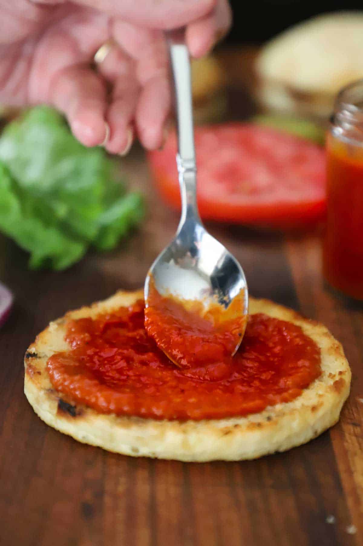 A person using a small spoon to smear easy homemade ketchup all over the top of a toasted hamburger bun on a wooden cutting board.