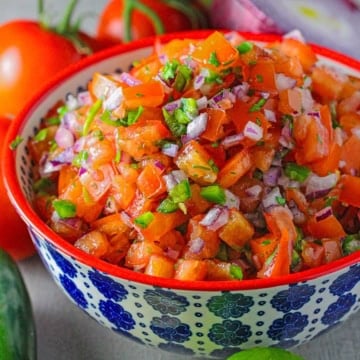 A close-up view of a colorful bowl of filled with a pile freshly made pico de gallo with chopped tomatoes, red onion, jalapeño, and cilantro.