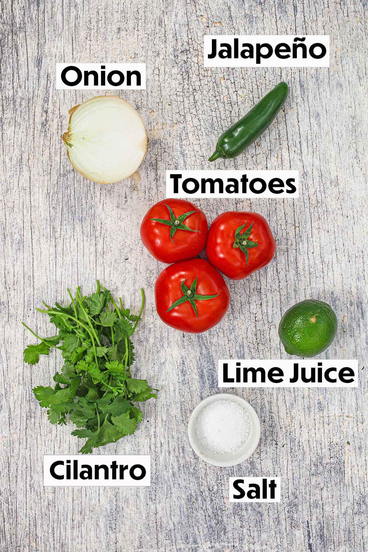An arrangement of ingredients for fresh pico de gallo on a grey wooden background including onion, jalapeño, tomatoes, cilantro, lime, and salt. 