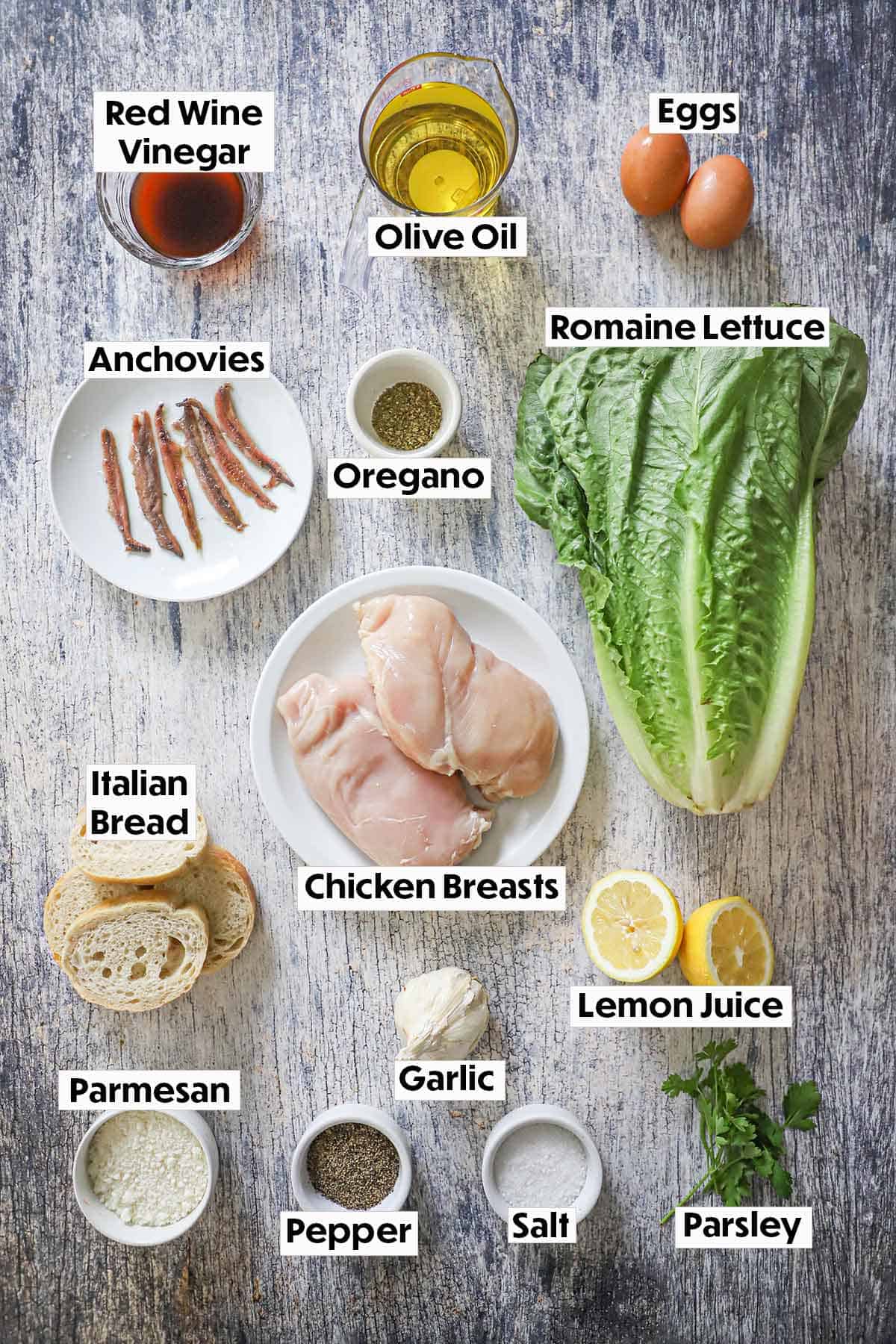 An arrangement of ingredients for grilled chicken Caesar salad on a grey wooden background including romaine lettuce, chicken breasts, eggs, lemons, anchovies, bread, Parmesan cheese, and seasonings.