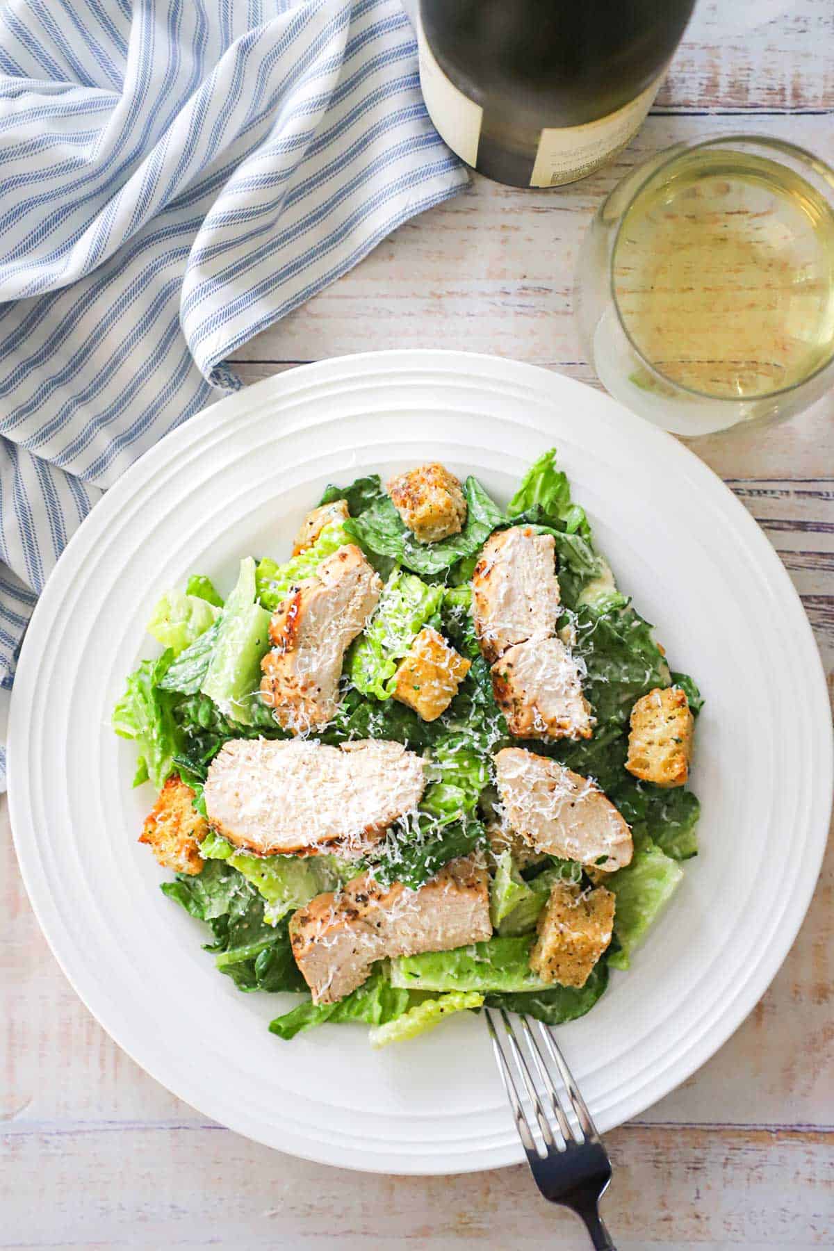 A white dinner plate filled with grilled chicken Caesar salad with a glass of white wine next to it.