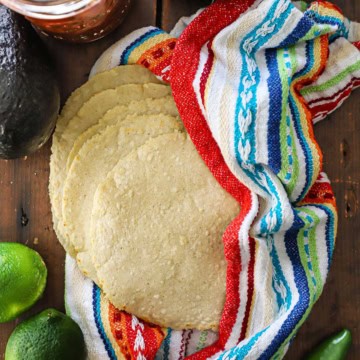 A spread-out stack of homemade corn tortillas that are nestled into a colorful napkin sitting on a wooden background and surround by whole limes, a jalapeño peppers, and an avocado.