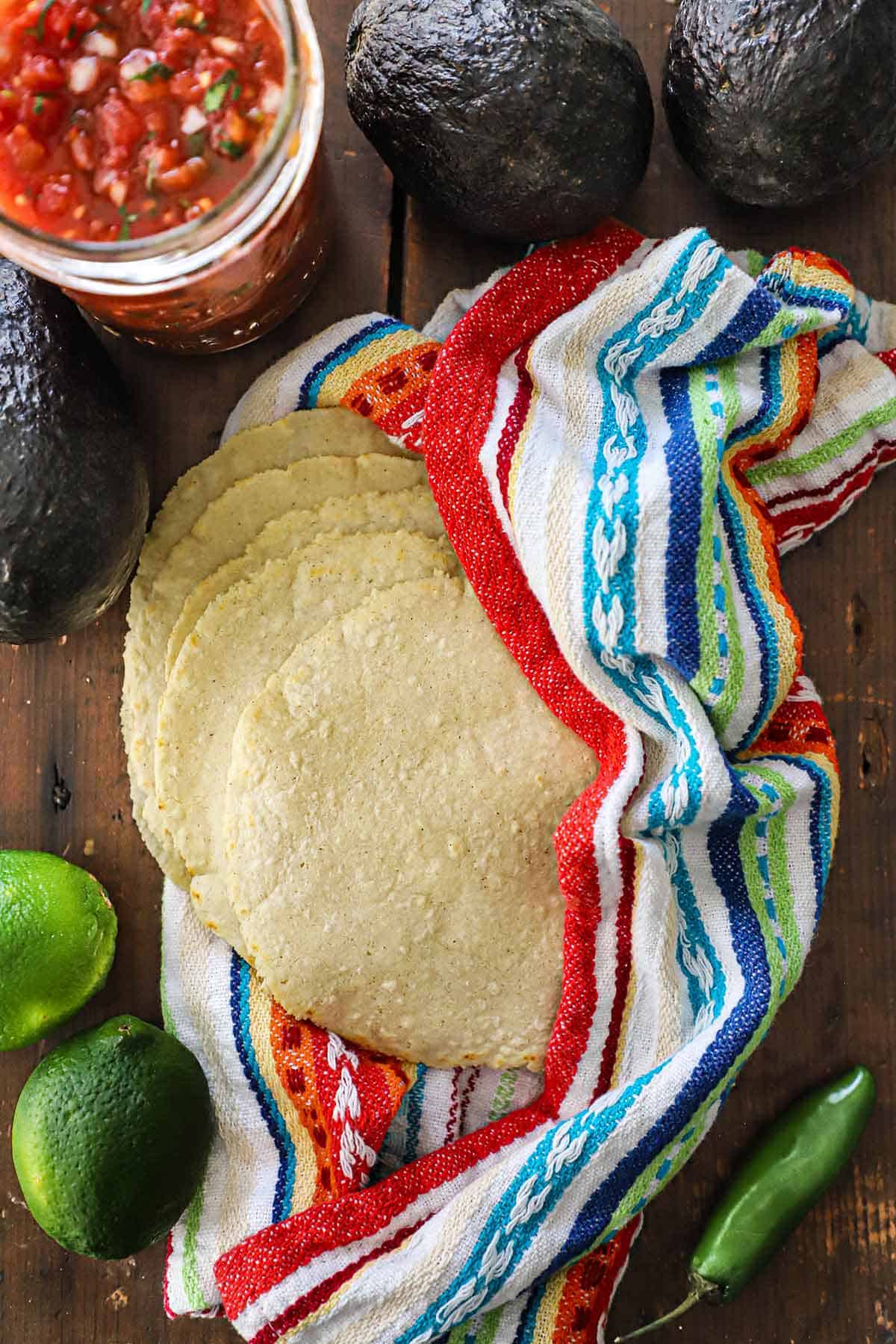 A spread-out stack of homemade corn tortillas that are nestled into a colorful napkin sitting on a wooden background and surround by whole limes, a jalapeño pepper, avocados, and a jar of salsa.