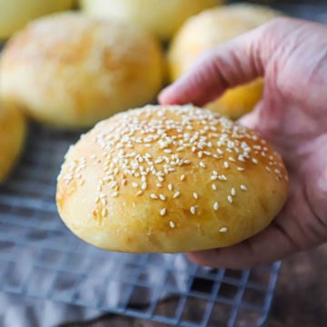 A person holding a homemade hamburger bun that is topped with sesame seeds with his hand over a baking rack filled with more buns.