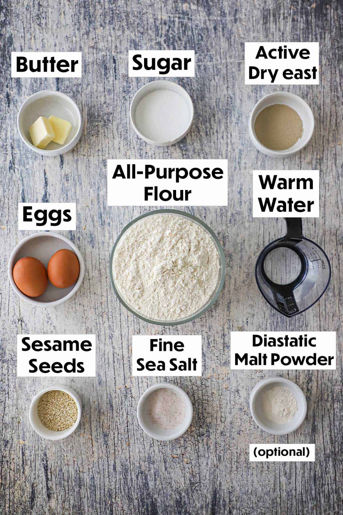 An arrangement of ingredients on a wooden grey background including flour, eggs, yeast, sugar, butter, malt powder, salt, and sesame seeds.