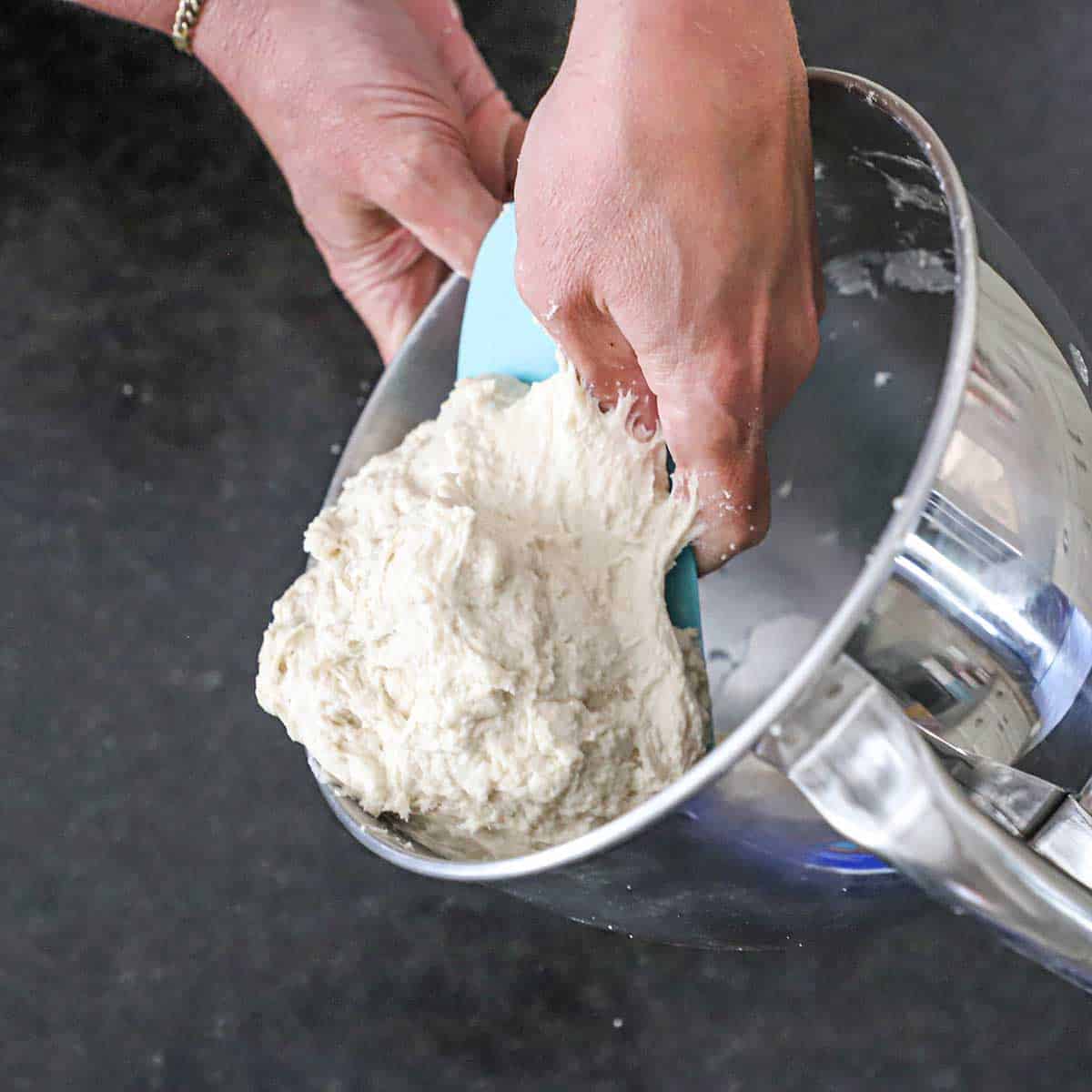 A person using a rubber dough scraper to remove New York-style pizza dough from the bowl of a stand mixer.