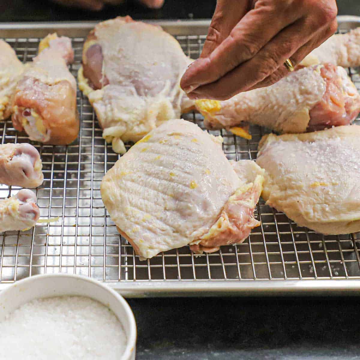 A person sprinkling salt over raw bone-in, skin on chicken pieces that are sitting on baking rack on a sheet pan.