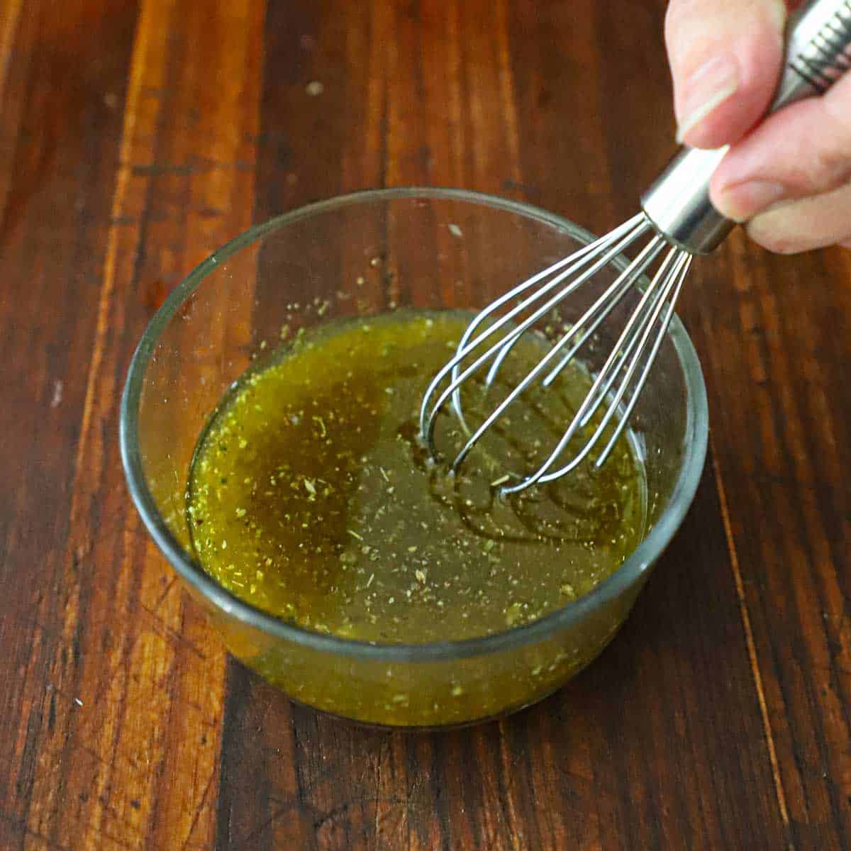 A person using a whisking together the ingredients for a marinade for grilled chicken in a small glass bowl on a wooden cutting board.
