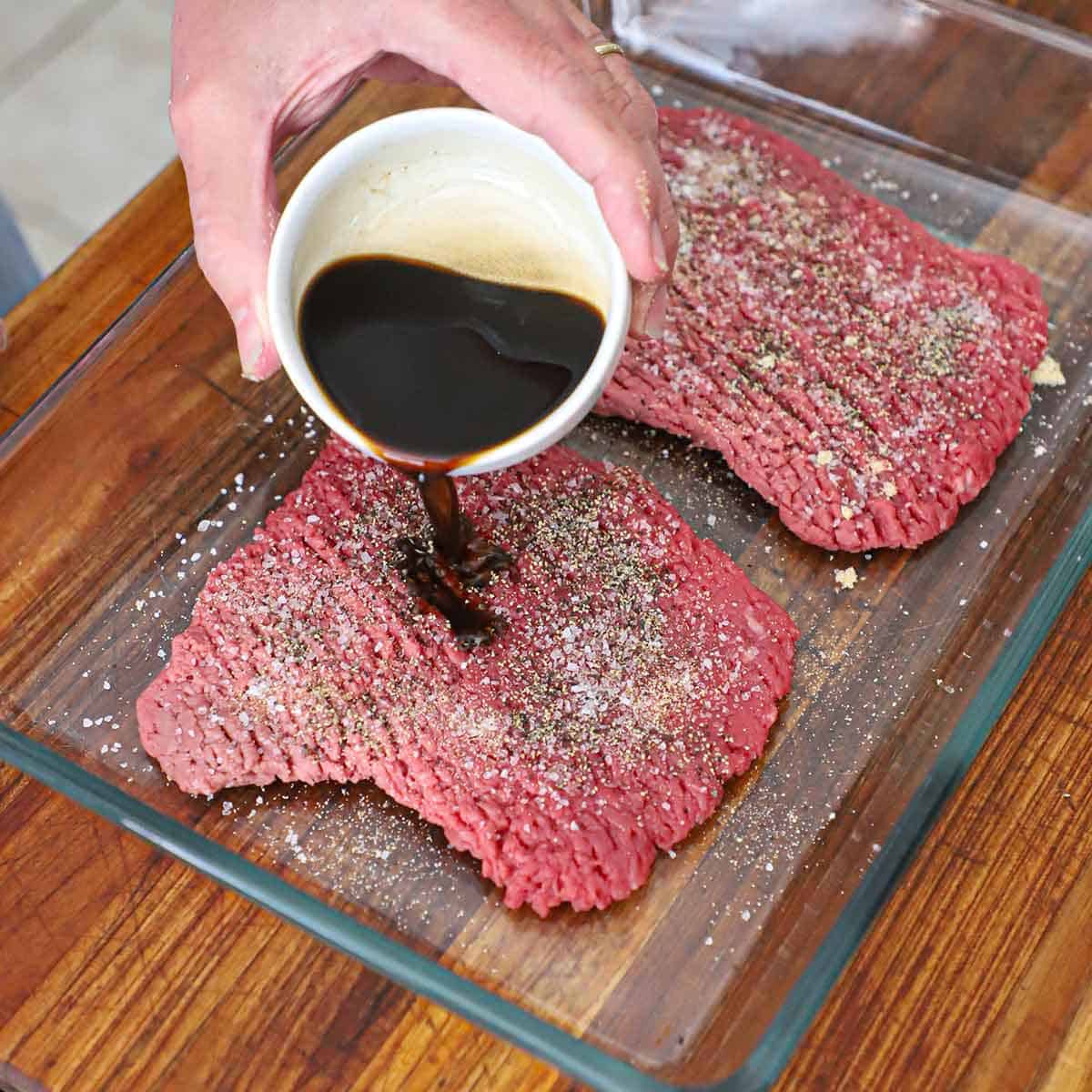 A person pouring Worcestershire sauce over two seasoned uncooked cube steaks in a glass baking dish.