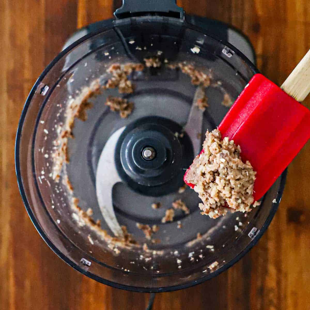 A person holding up a rubber red spatula with finely chopped anchovies and garlic over a the bowl food processor.