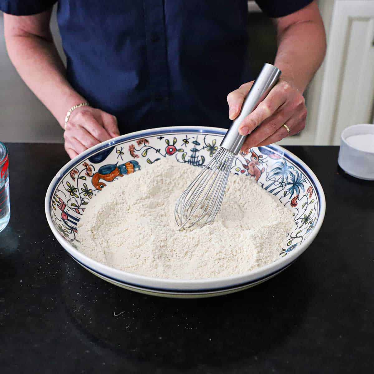 A person using a large metal whisk to combine finely-ground masa harina and salt in a large, colorful, shallow bowl in a black marble counter.