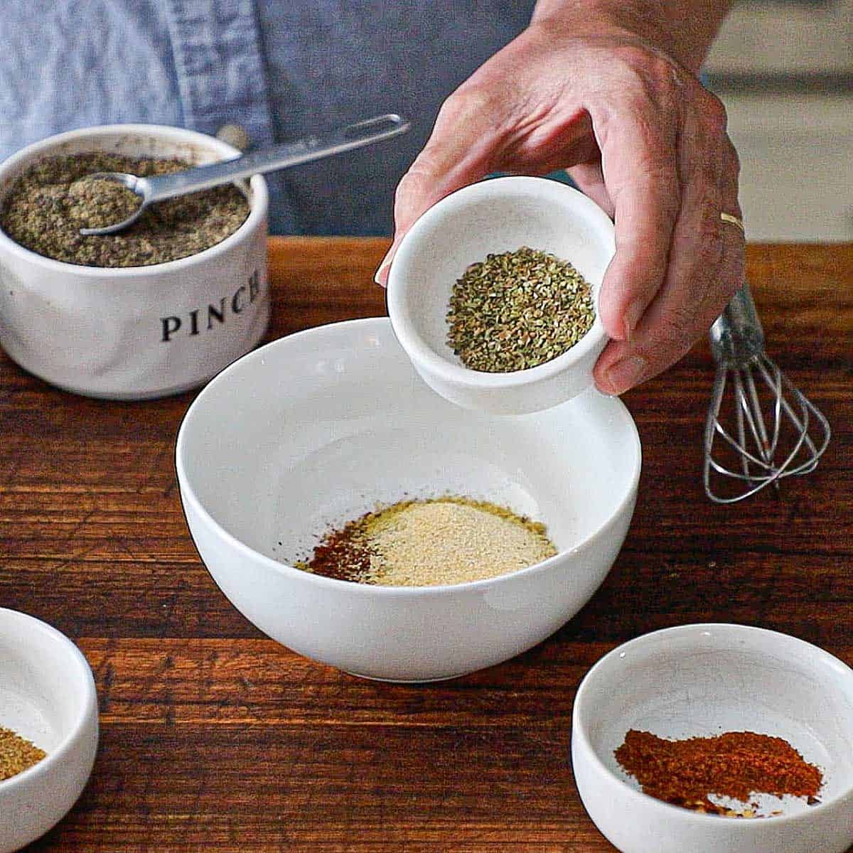 A person holding a small white bowl filled with dried oregano over another larger bowl filled with chili powder and other seasonings.