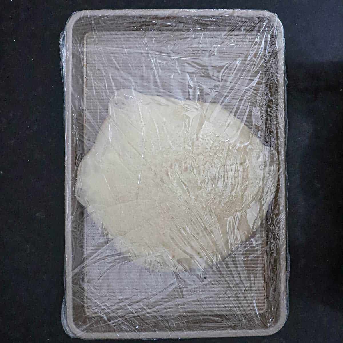 An expanded ball of New York-style pizza dough on a gold sheet pan that has been twice wrapped in plastic wrapy.