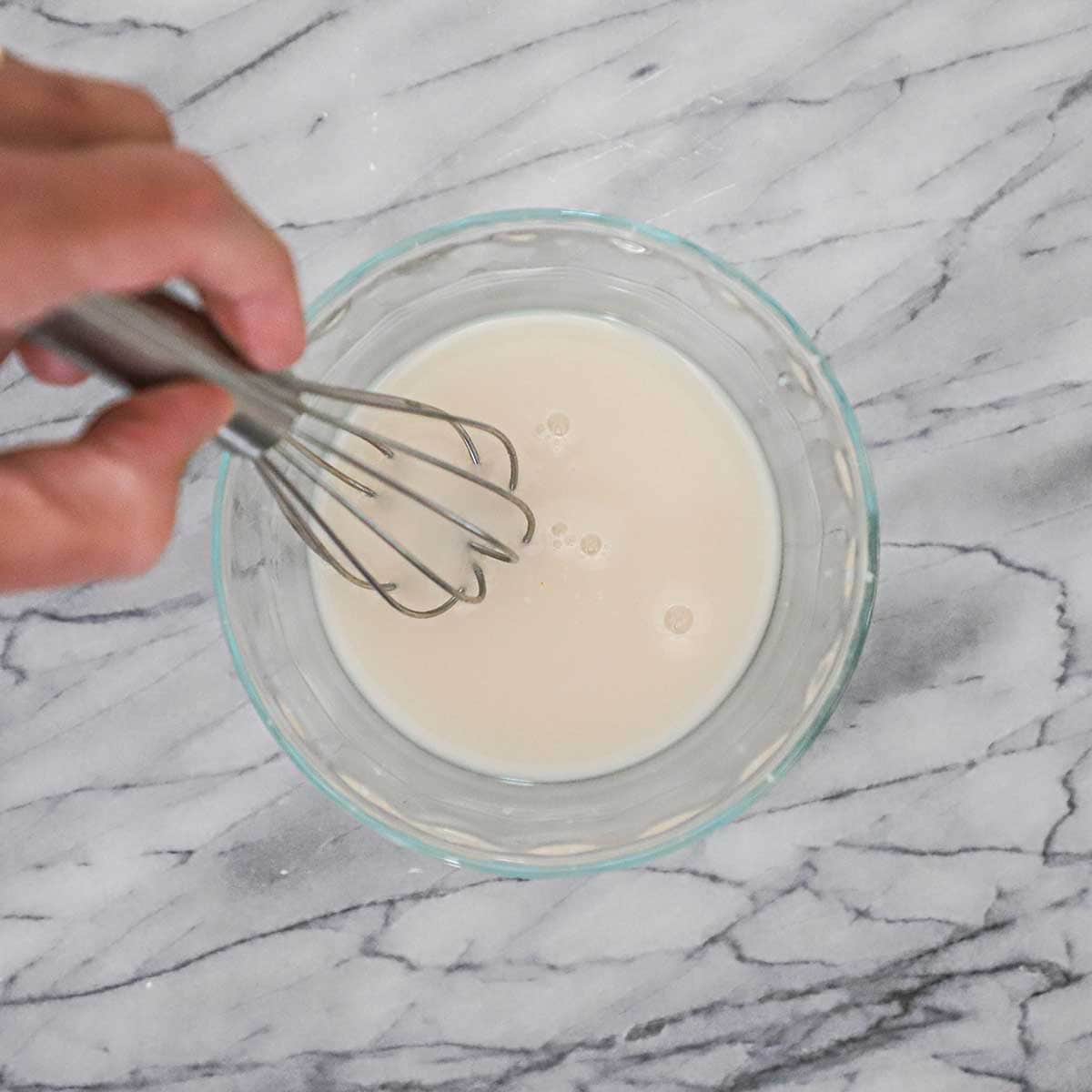 A person using a small whisk to activate active dry yeast in a small bowl of water on a marble counter.