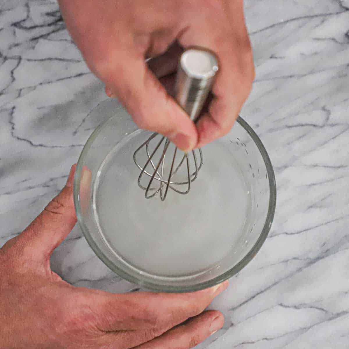 A person using a small whisk to activate a small amount of active yeast in a small glass bowl.