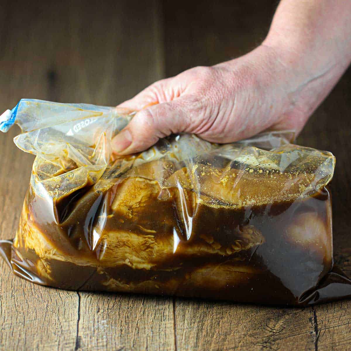 A person using his hand to squeeze a plastic freezer bag that is filled with skinless chicken breasts in a dark brown fajita marinade.