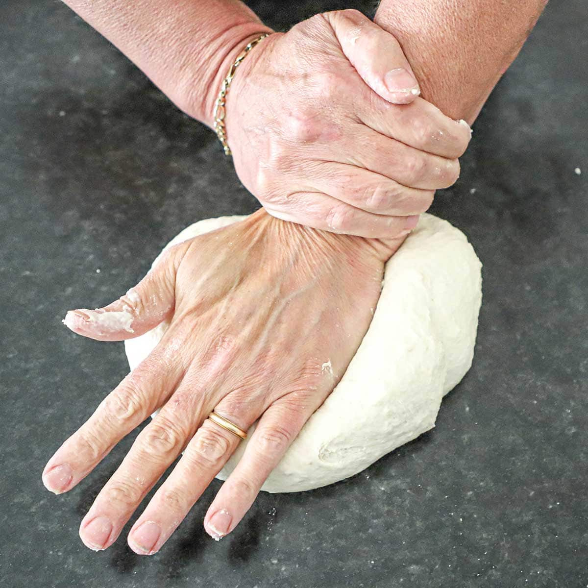 A person using his hands to knead New York-style pizza dough on a black marbel counter.
