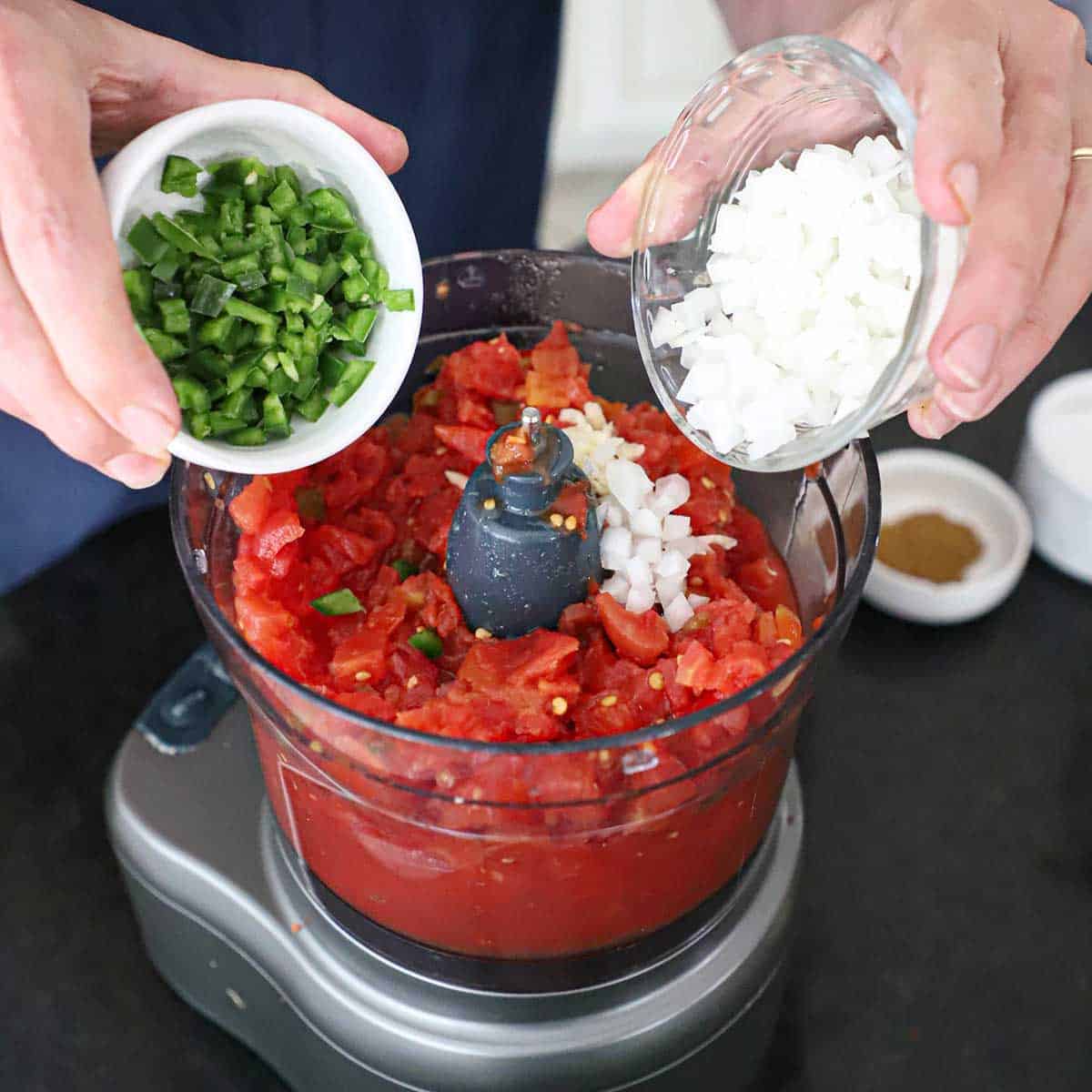 A person adding chopped jalapeño and chopped white onion from small bowl into a food processor that is filled with chopped Ro-tel tomatoes and whole canned tomatoes.