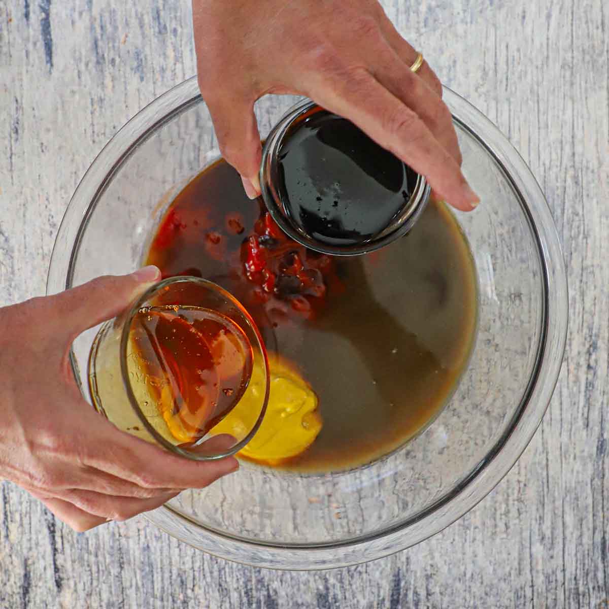 A person holding a small bowl of molasses in one hand and the other hand holding a small bowl of honey over a large glass bowl filled ketchup, mustard and other ingredients for homemade BBQ sauce.