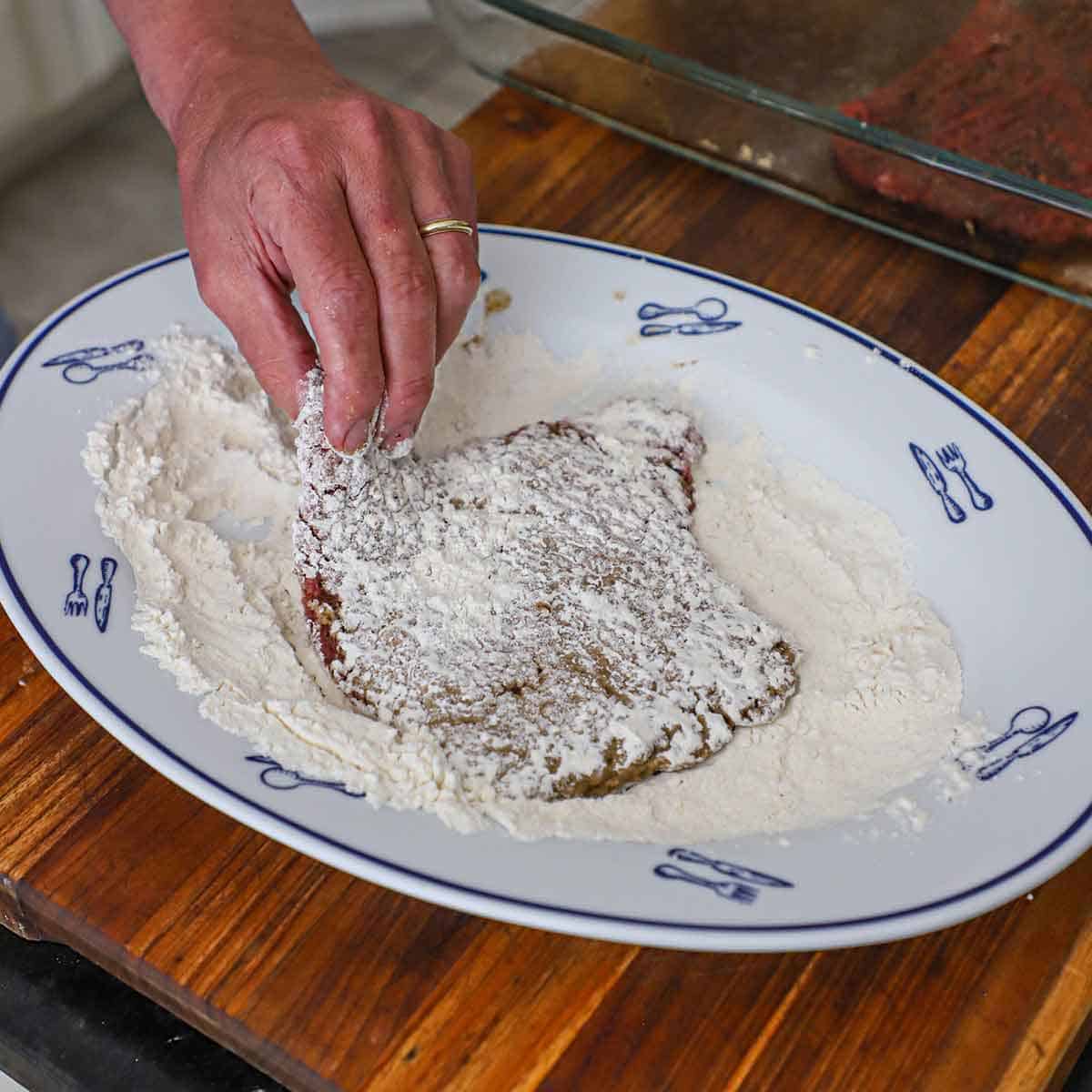 A person dredging a seasoned cube steak through flour on a large oval platter sitting on a wooden cutting board.