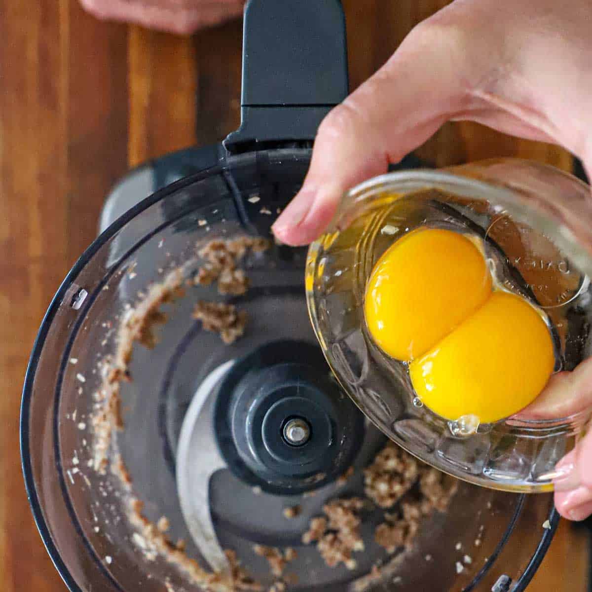 A person transferring two egg yolks into the bowl of a food processor that is filled with finely chopped anchovies and garlic.