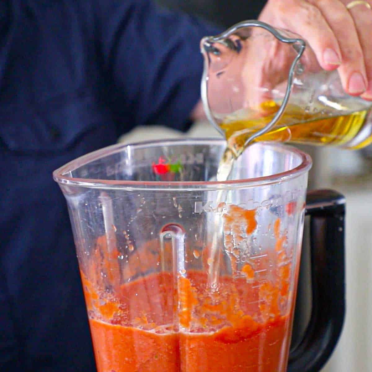 A person pouring apple cider vinegar from a glass measuring cup into a blender filled with puréed tomatoes and vegetables.