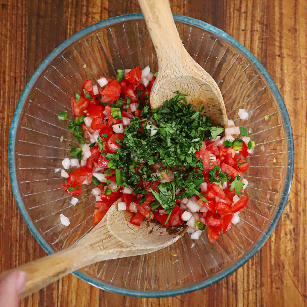 A glass bowl filled with chopped tomatoes, onions, jalapeño, and chopped cilantro with two wooden spoons being used to toss it all together.