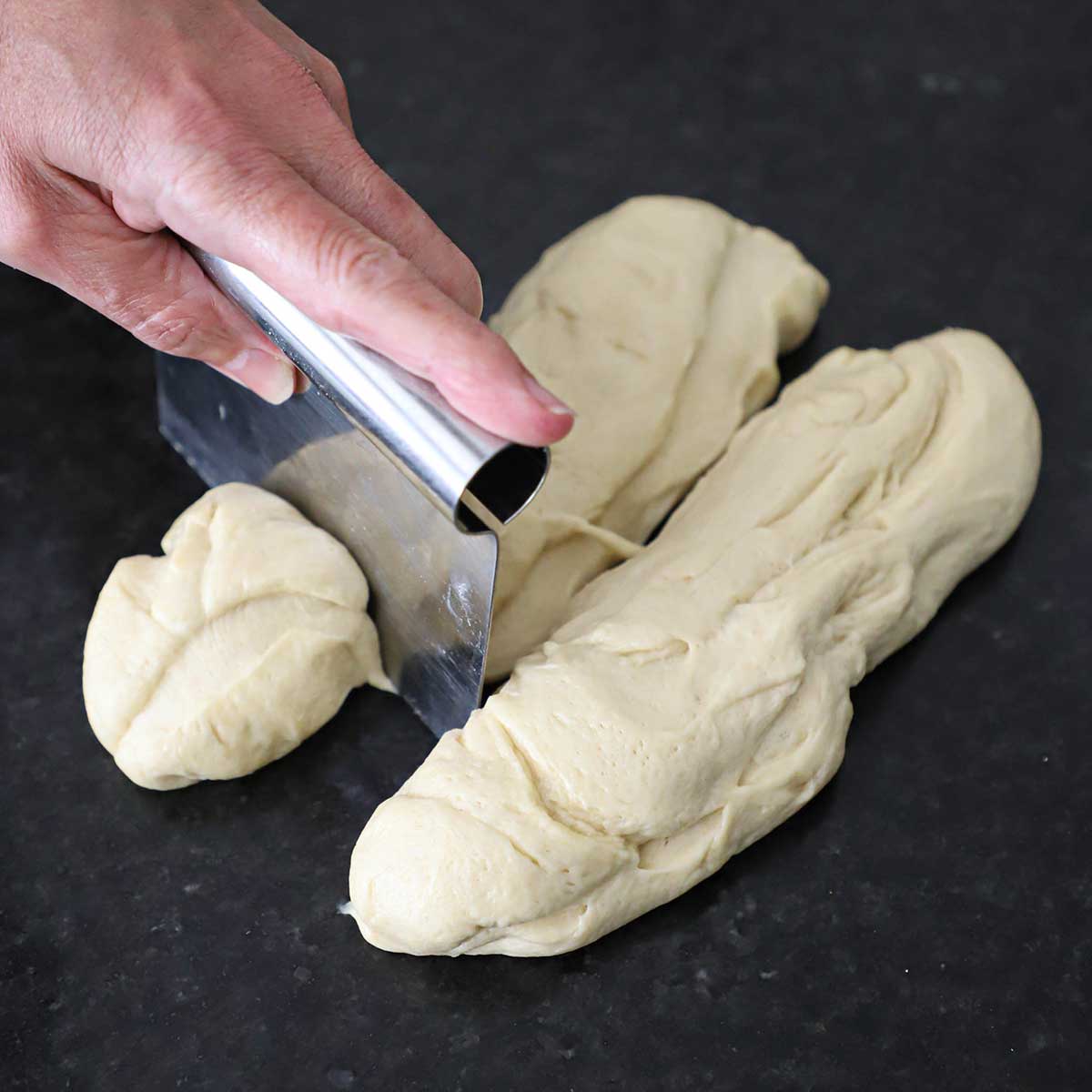 A person using a metal bench scraper to cut two logs of hamburger bun dough into smaller portions on a black marble counter.