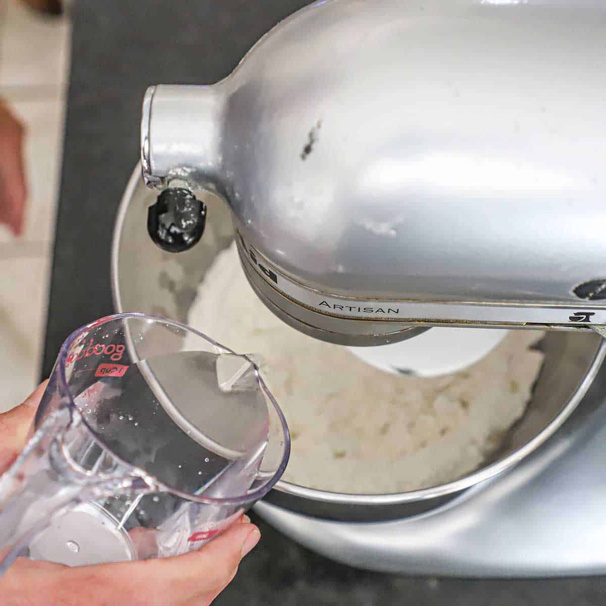A person pouring water into a the bowl of a stand mixer that is filled with the ingredients for New York-style pizza dough.
