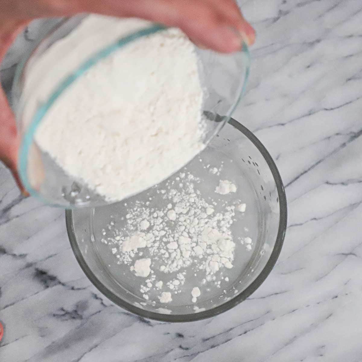 A person dumping high-gluten flour into a small glass bowl that is filled with a water and active dry yeast mixture.