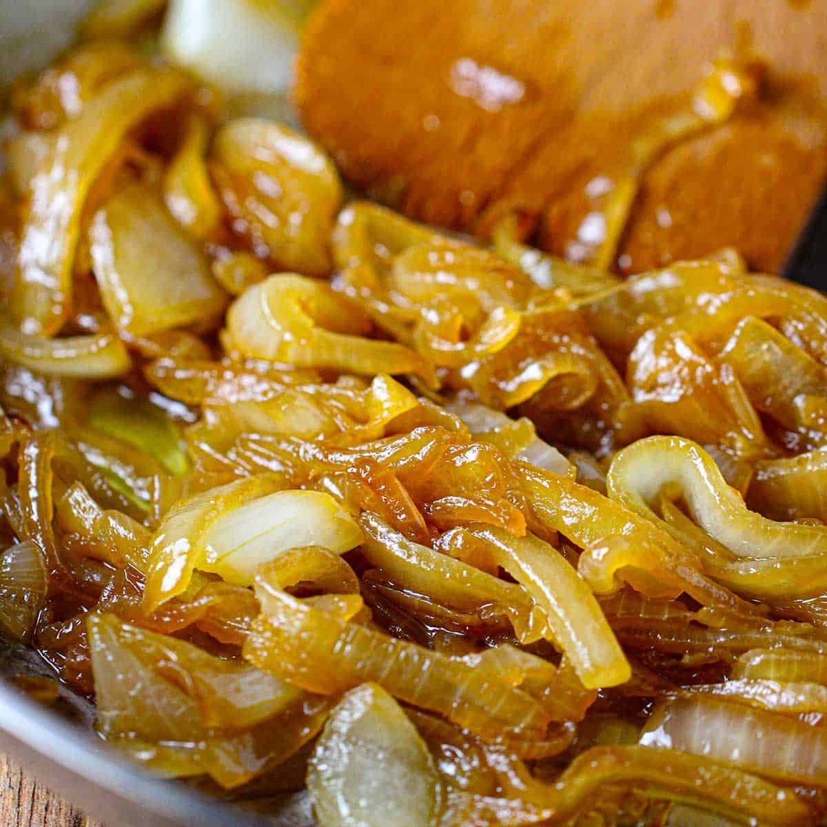 A close-up view of thinly sliced onions that have been caramelized in a skillet with a wooden spatula.
