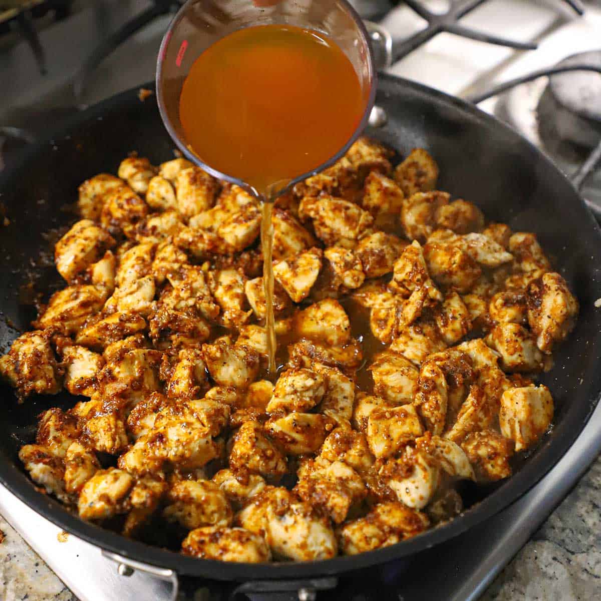 A person pouring chicken broth from a glass measuring cup into a skillet filled with chunks of cooked chicken breast simmering in oil and taco seasoning.
