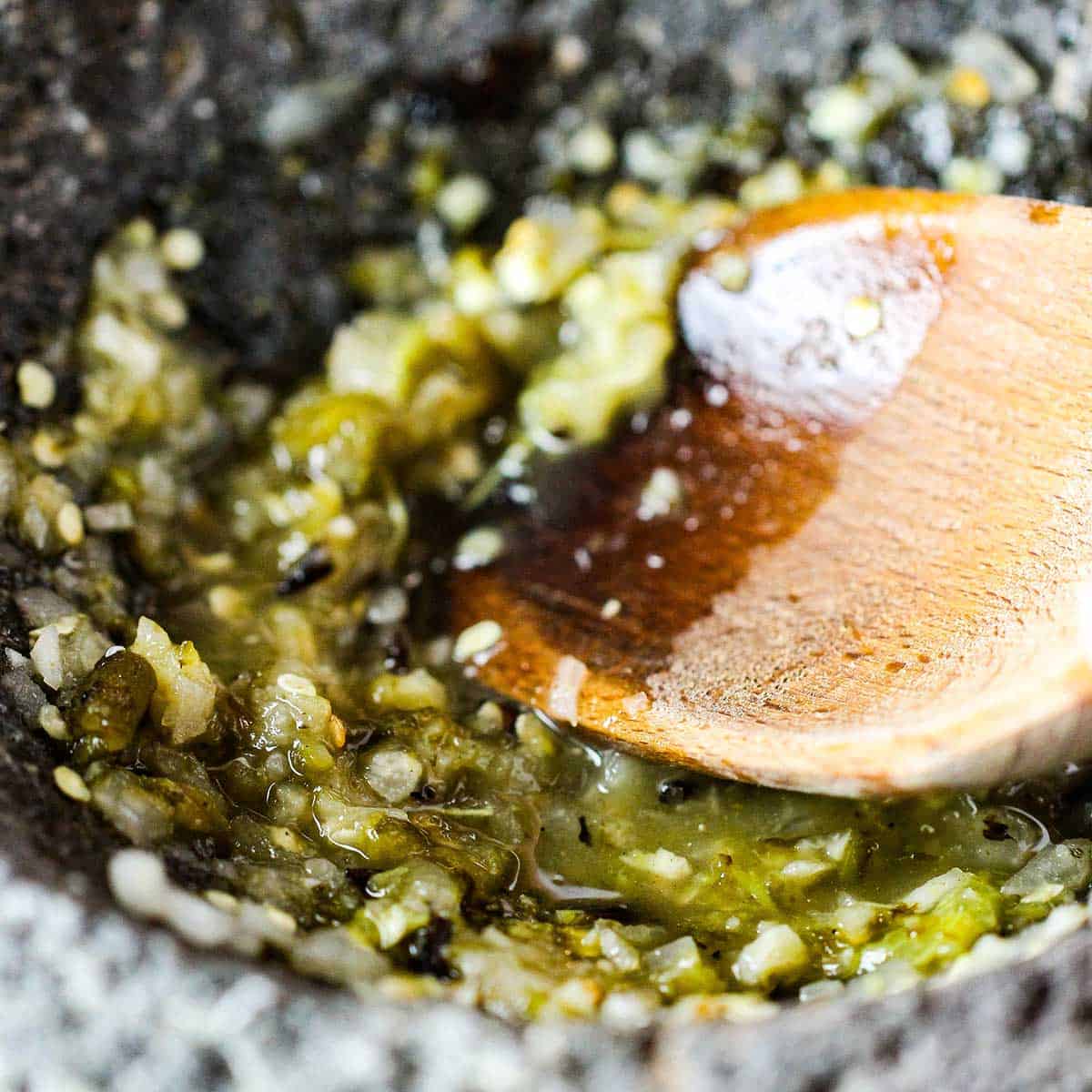 A close-up view of a wooden spoon muddling roasted peppers, onions, and garlic in the bottom of a molcajete.