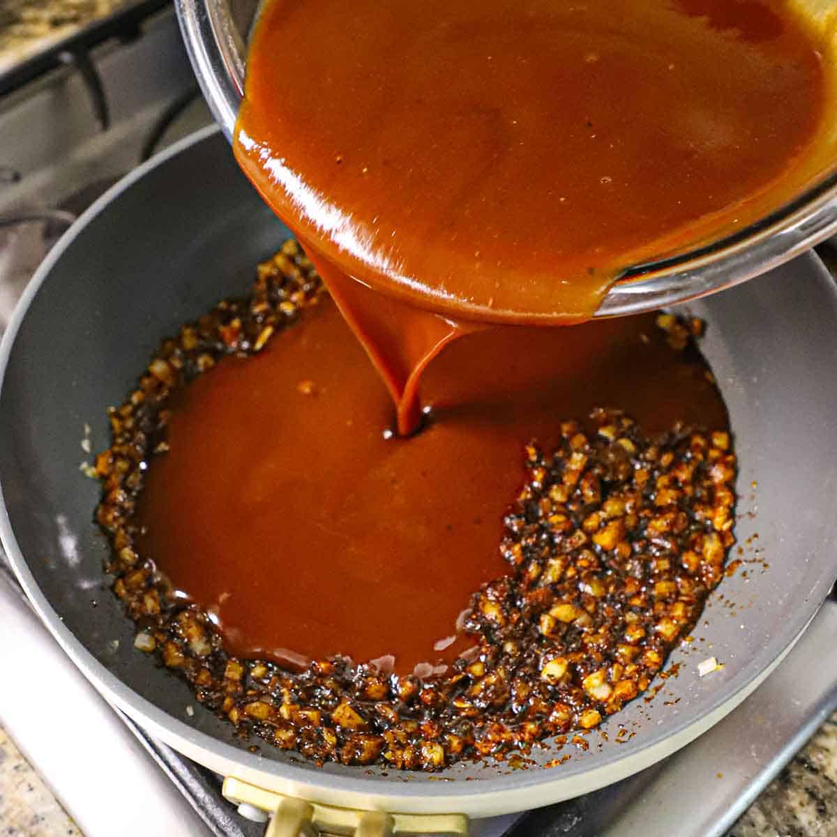 A person pouring a brown sauce into a large skillet filled with sautéed onions and spices.