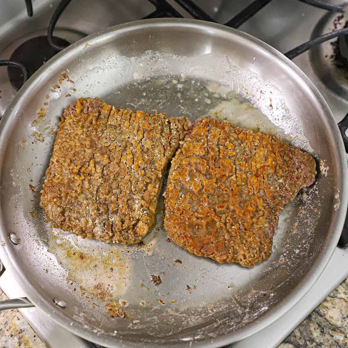 Two browned cubed steaks resting in a stainless steel skillet sitting on a gas stove.