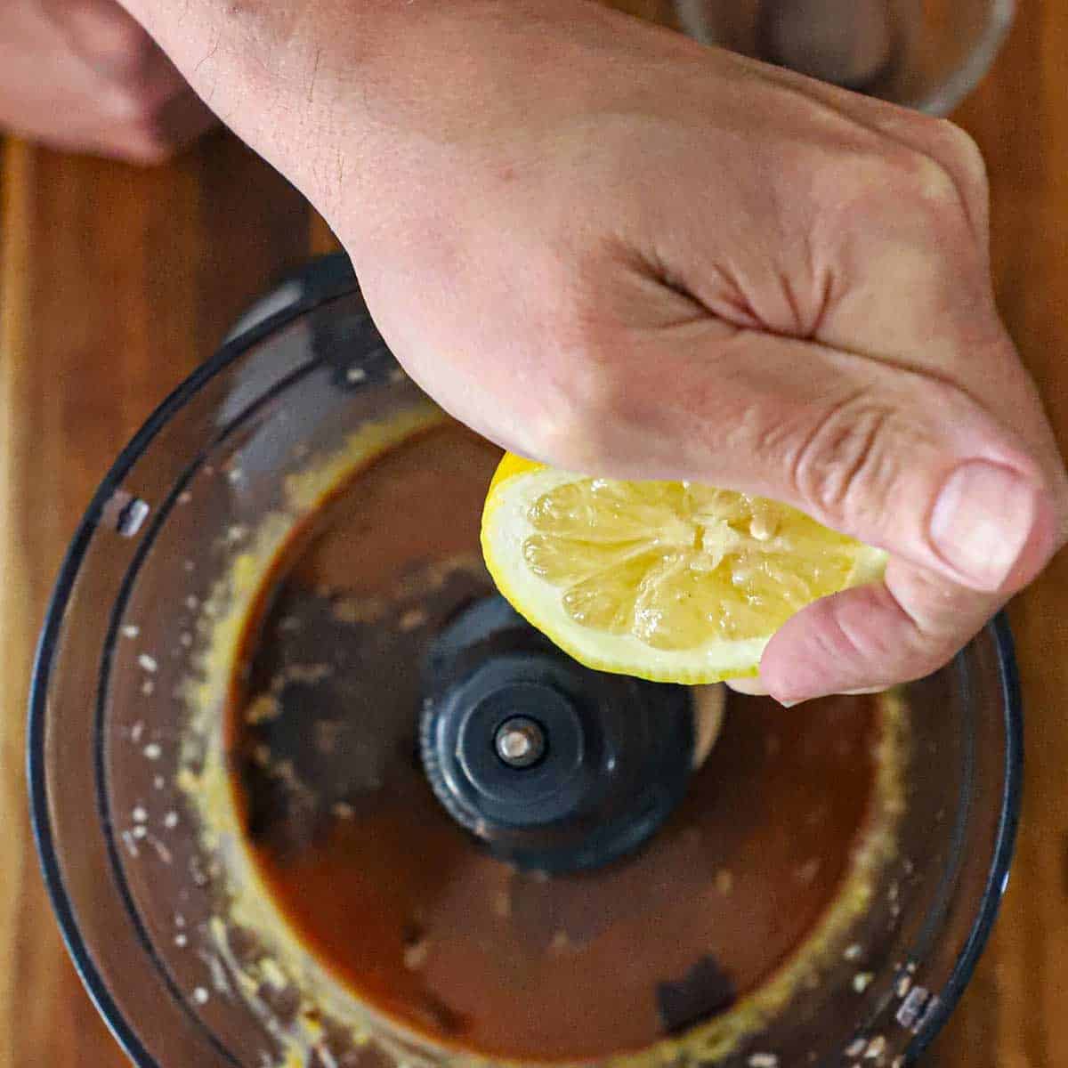 A person squeezing half of a lemon into the bucket of a food processor that is filled with puréed egg yolks mixed with chopped anchovies and garlic.