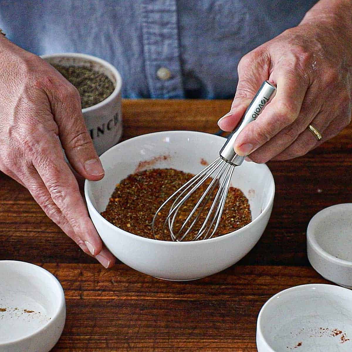 A person using a small whisk to combine seasonings in a white bowl to create homemade taco seasoning.