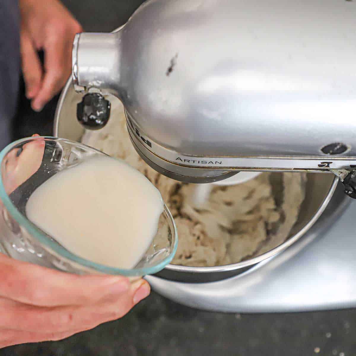 A person pouring a water and active dry yeast mixture into a bowl of a stand mixer that is filled with a flour and water mixture.